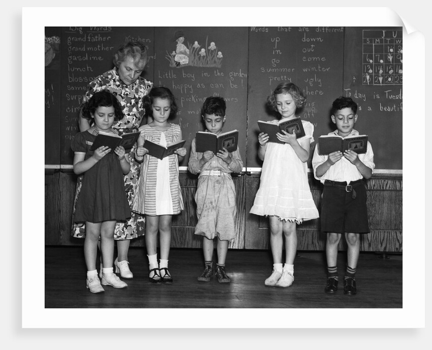 1930s line-up of 5 elementary school students in front of blackboard reading books with teacher looking on by Corbis