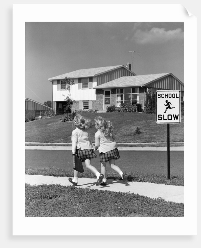 1950s back view of twin girls in plaid skirts & cardigans holding book bags running past school slow sign by Anonymous