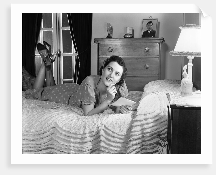 1940s fully dressed smiling woman laying on her bed with her legs up reading a letter in her bedroom by Anonymous