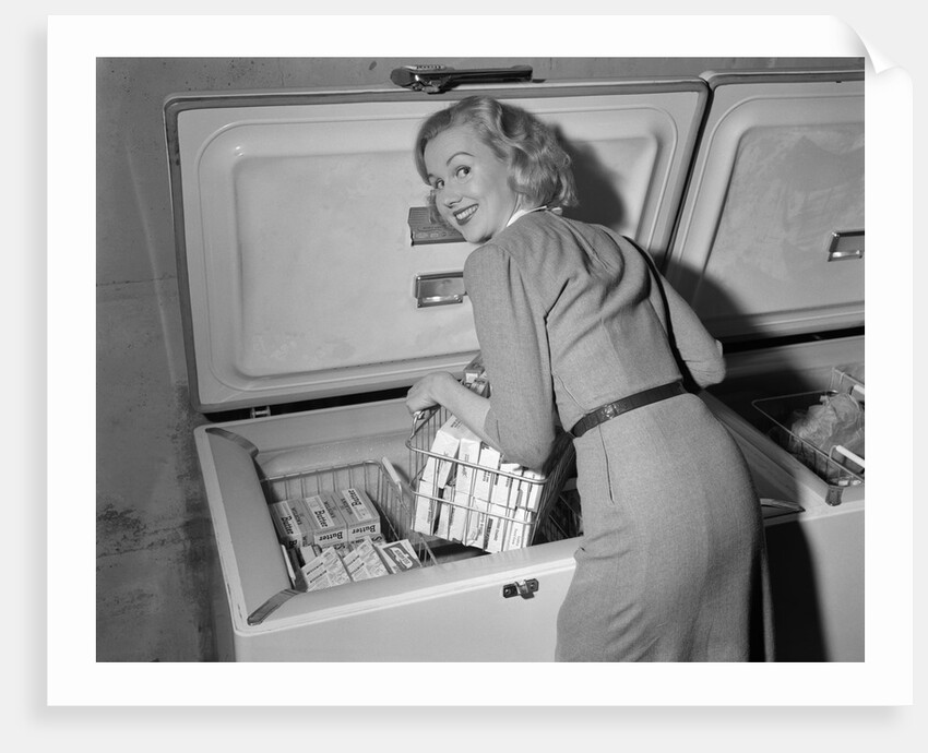 1950s blond woman lifting wire basket food items from a deep freezer looking at camera over her shoulder by Anonymous
