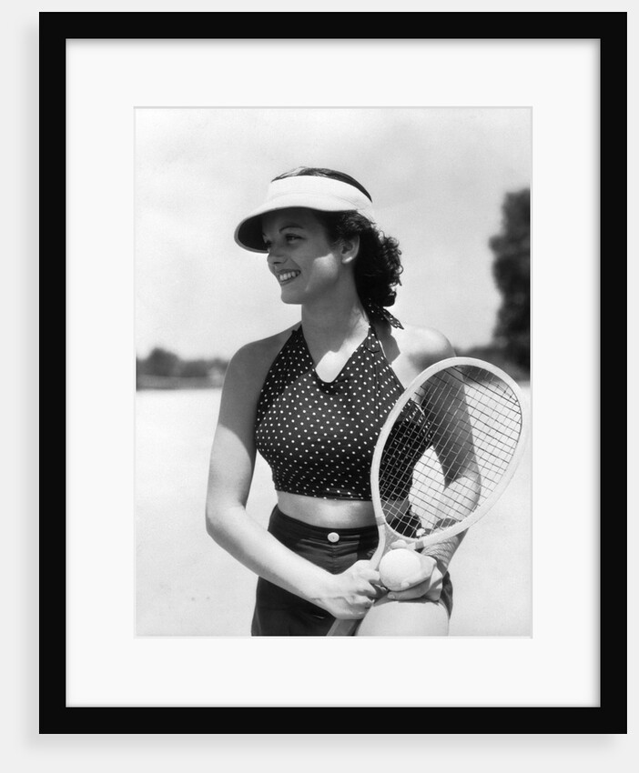 1930s woman in polka dot halter top shorts & sun visor holding a tennis ball & racket by Anonymous