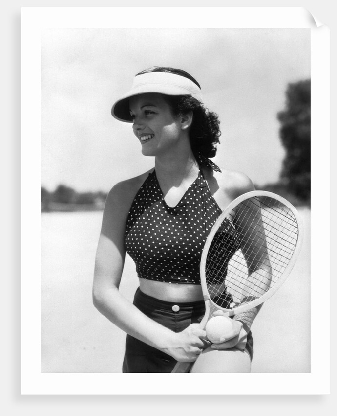 1930s woman in polka dot halter top shorts & sun visor holding a tennis ball & racket by Anonymous