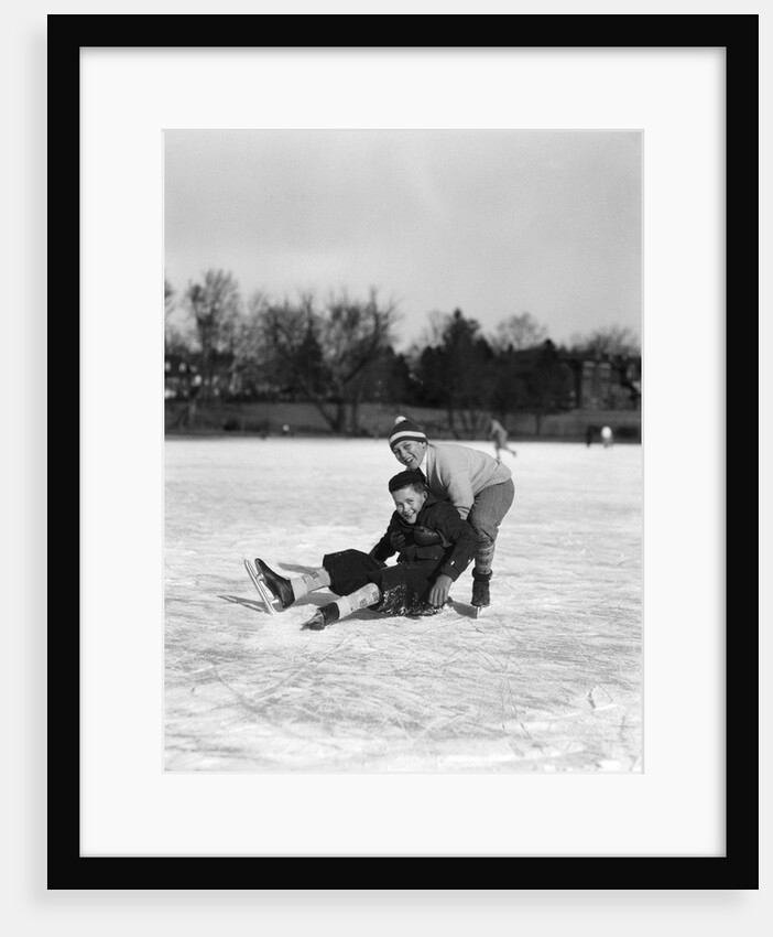 1920s 1930s two smiling boys looking at camera ice skating one boy fallen other picking him up by Anonymous