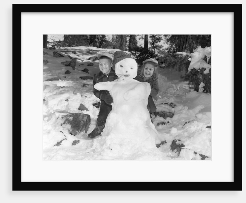 1950s 1960s smiling boy and girl building a snowman together in snowy woods by Anonymous