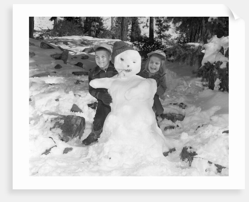 1950s 1960s smiling boy and girl building a snowman together in snowy woods by Anonymous