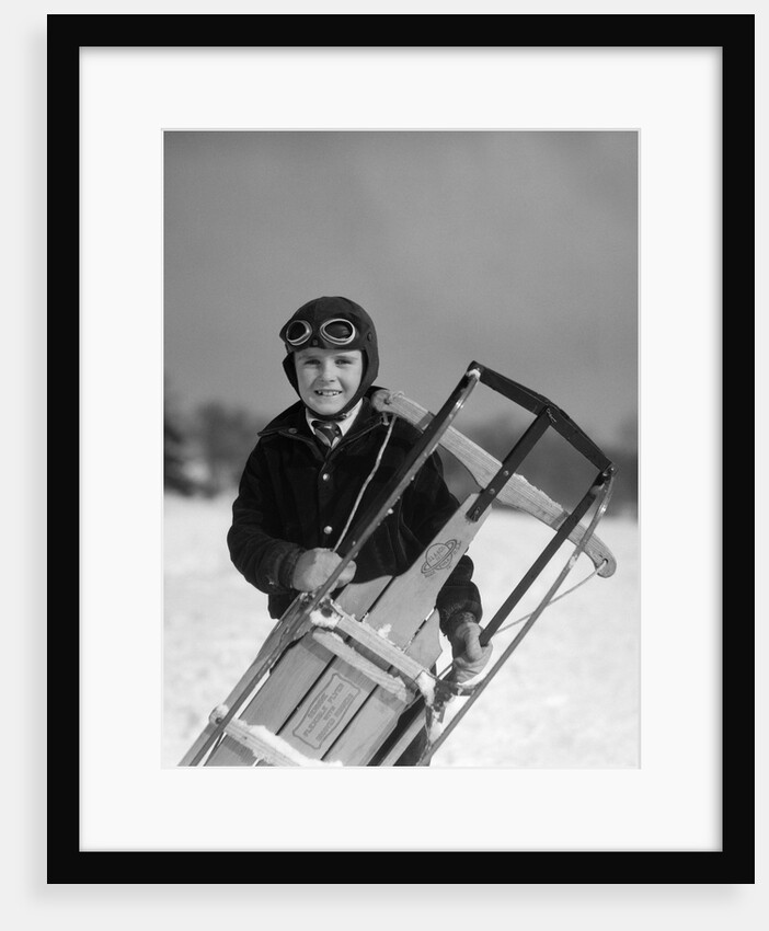 1920s 1930s smiling boy wearing aviator goggles leather flying helmet holding sled standing in snow field looking at camera by Anonymous