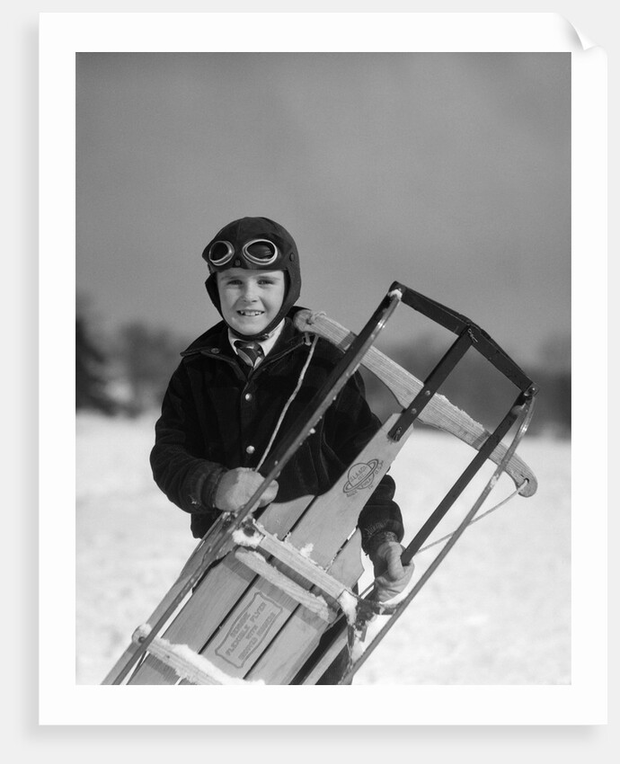 1920s 1930s smiling boy wearing aviator goggles leather flying helmet holding sled standing in snow field looking at camera by Anonymous