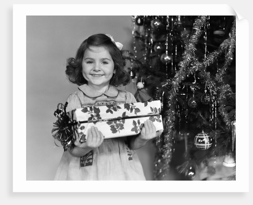 1940s little girl standing near christmas tree holding a wrapped present smiling looking at camera by Anonymous