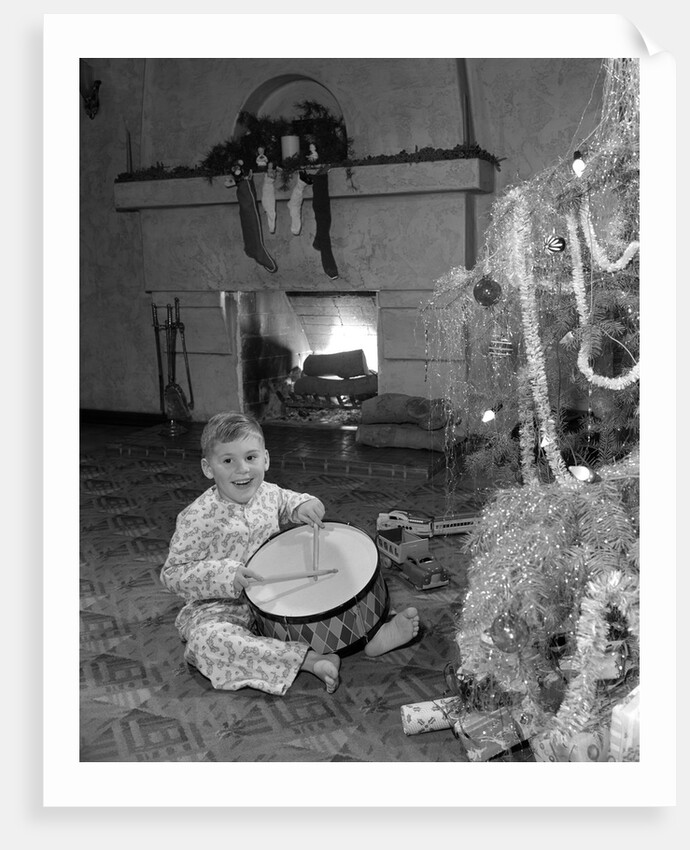 1950s little boy playing toy drum by christmas tree and fireplace looking at camera by Anonymous