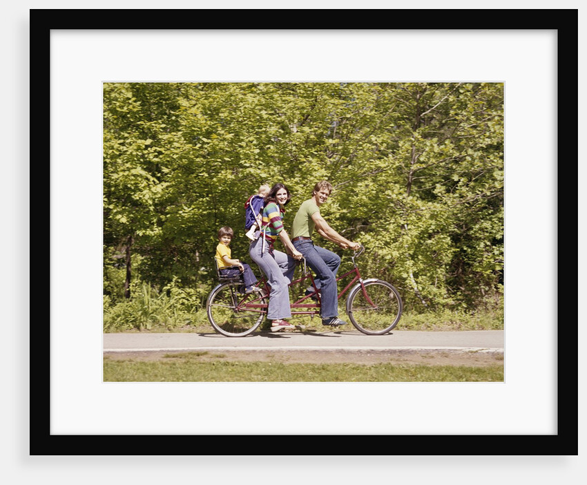 1970s Family On Tandem Bicycle Mother Father Son & Baby Daughter In Backpack Wearing Bellbottom Blue Jeans Looking At Camera by Anonymous