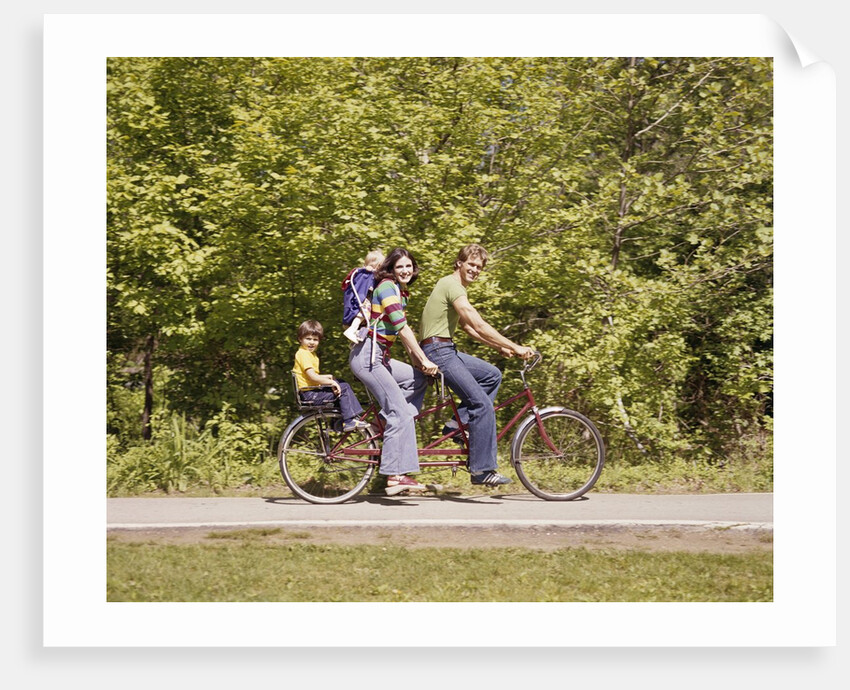 1970s Family On Tandem Bicycle Mother Father Son & Baby Daughter In Backpack Wearing Bellbottom Blue Jeans Looking At Camera by Anonymous
