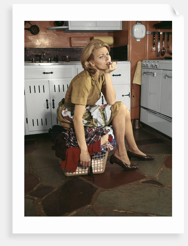 1960s Tired Housewife Sitting On Top Of Laundry Basket In Kitchen by Anonymous
