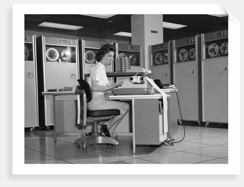 1960s Woman In Mainframe Computer Room Surrounded By Many Data Tape Drives Sitting At Desk Typing On Typewriter Indoor by Anonymous