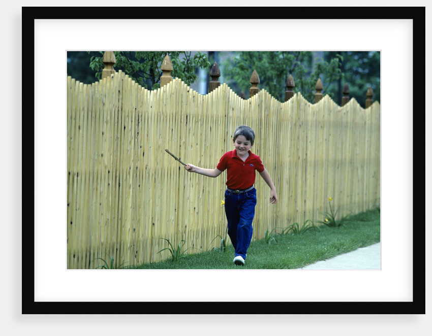 1980s Smiling Boy Running Along Sidewalk Rattling Stick On Tall Picket Fence Making Noise by Anonymous