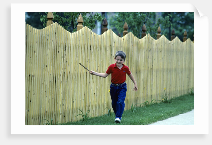 1980s Smiling Boy Running Along Sidewalk Rattling Stick On Tall Picket Fence Making Noise by Anonymous