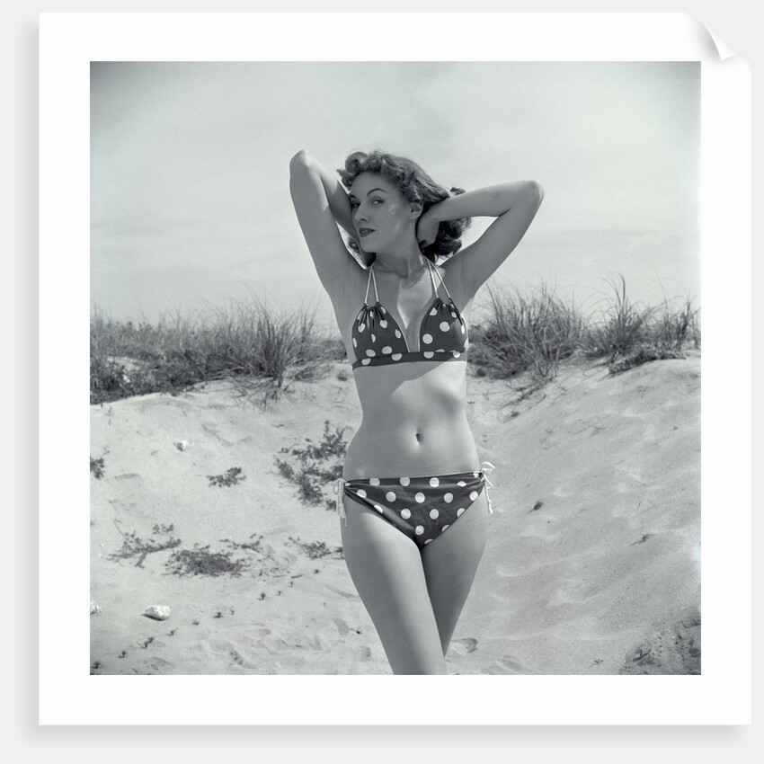 1950s Brunette Bathing Beauty In Polka Dot Bikini Standing In Sand With Hands Behind Head Looking At Camera by Anonymous