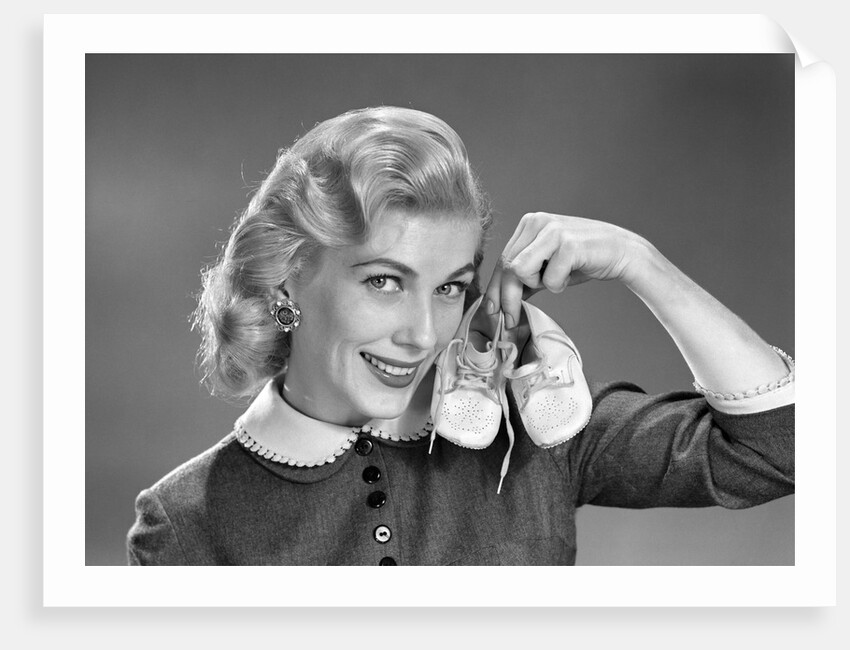 1950s 1960s Smiling Blond Woman Communicating That She Is Mother To Be By Holding Up A Pair Of Baby Shoes Looking At Camera by Anonymous
