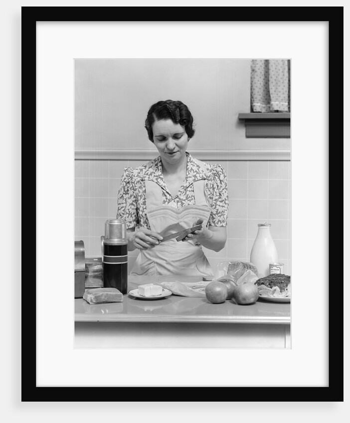 1930s Woman Housewife In Kitchen Wearing Apron Making Sandwich Packing Metal Lunch Box Thermos Apples Bread Milk On Counter by Anonymous