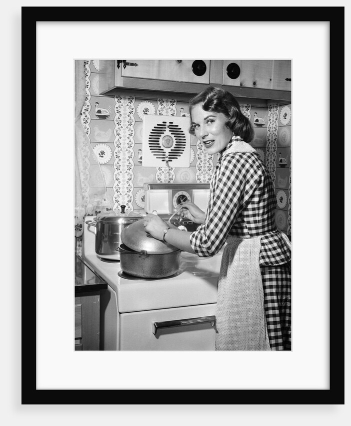 1950s Housewife Wearing Checkered Dress Standing In Kitchen Stirring Pot On Stove Looking Over Shoulder by Anonymous