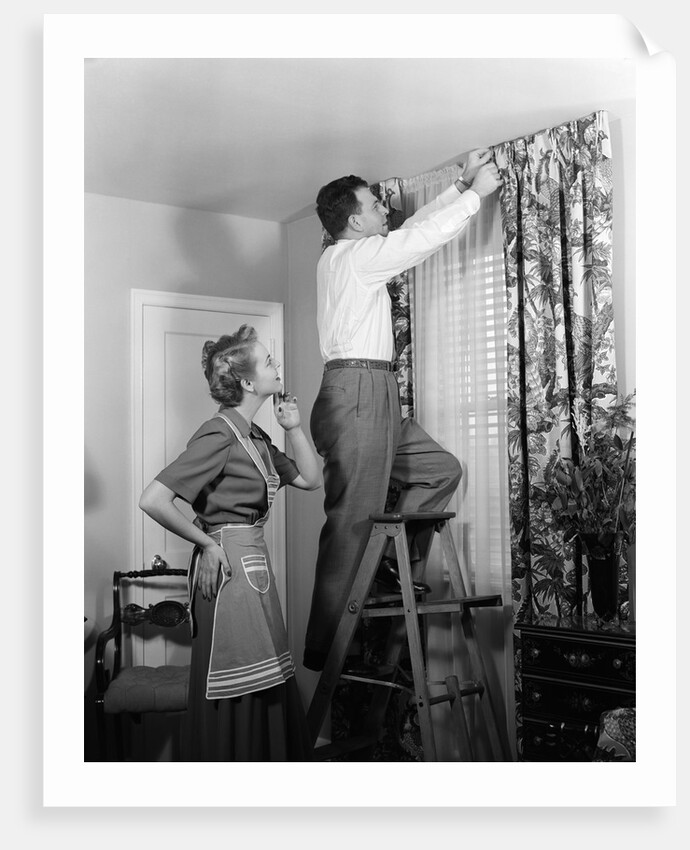 1950s Skeptical Wife In Apron Fingers Pressing Chin Watching Husband On Ladder Hanging New Jungle Motif Drapes In Living Room by Anonymous