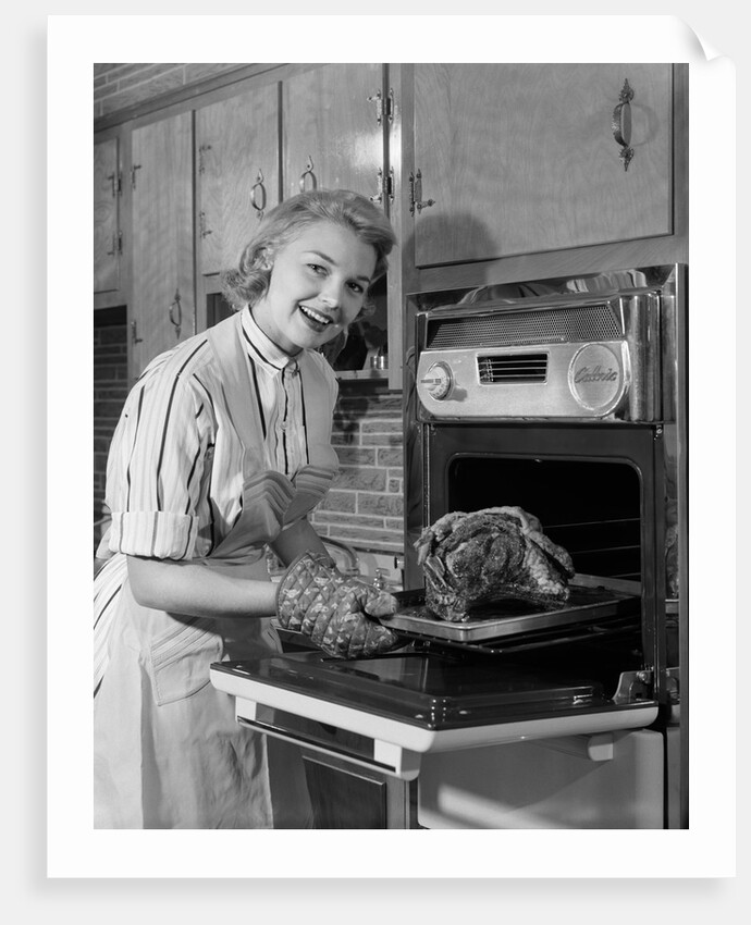 1950s Smiling Woman Housewife Wearing Apron Taking Large Roast Beef From Electric Oven In Kitchen Looking At Camera by Anonymous