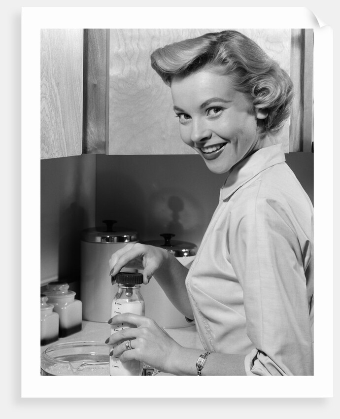 1950s Smiling Blonds Woman Mother Warming A Baby Bottle Of Milk In Kitchen Looking At Camera by Anonymous