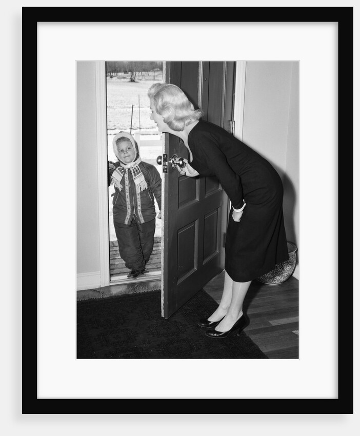 1950s Blonde Mom Opening Front Door To Let In Child Dressed Winter Snow Suit Scarf Hat Can See Snow Scene Through Door by Anonymous
