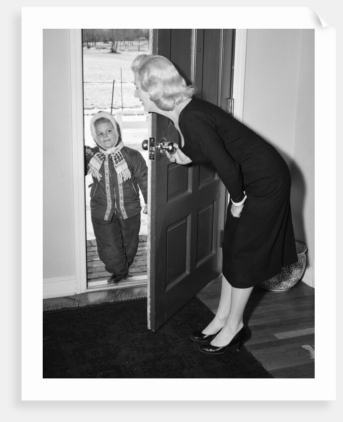 1950s Blonde Mom Opening Front Door To Let In Child Dressed Winter Snow Suit Scarf Hat Can See Snow Scene Through Door by Anonymous