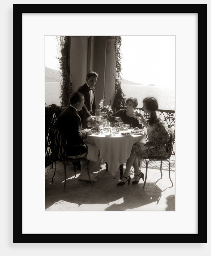 1920s group eating on balcony with waiter serving wine by Anonymous