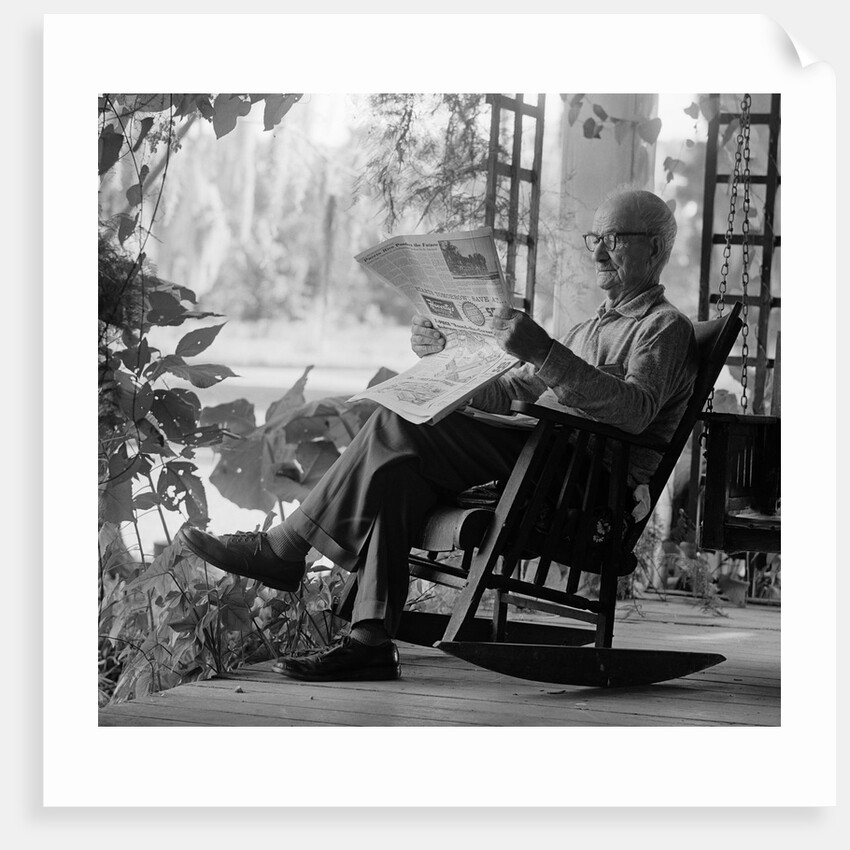 1970s elderly man reading newspaper on porch in a rocker by Anonymous