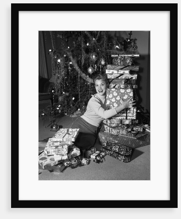1950s portrait of young woman holding stack of presents next to a Christmas tree by Anonymous