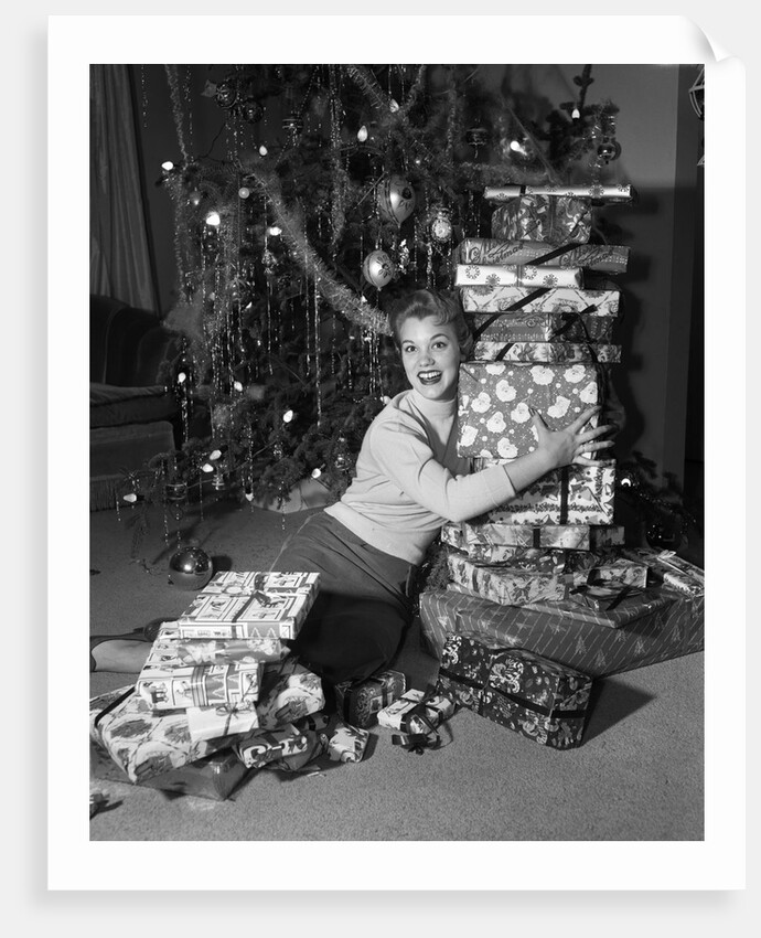 1950s portrait of young woman holding stack of presents next to a Christmas tree by Anonymous