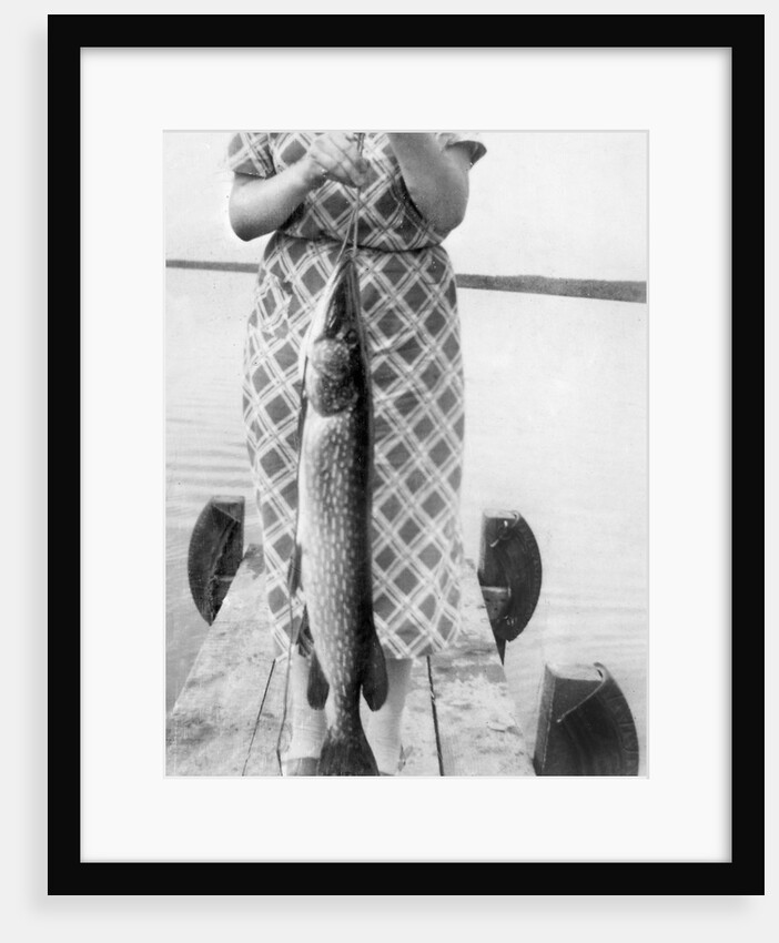 Woman holds on large Northern Pike on a lake pier, ca. 1950. by Anonymous