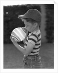 1950s Side View Of Boy In Baseball Cap and Striped T-Shirt Blowing Up Swirled Balloon by Anonymous