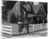 1950s Family Standing By White Fence Looking At Brick House by Anonymous
