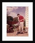 1960s Man Wearing Red Shirt Cooking Steak Outdoor On Backyard Grill by Anonymous