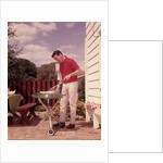 1960s Man Wearing Red Shirt Cooking Steak Outdoor On Backyard Grill by Anonymous
