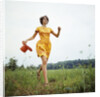 1970s Young Woman Wearing Colorful Print Dress Running In Field Carrying Red Straw Hat by Anonymous