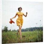 1970s Young Woman Wearing Colorful Print Dress Running In Field Carrying Red Straw Hat by Anonymous