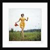 1970s Young Woman Wearing Colorful Print Dress Running In Field Carrying Red Straw Hat by Anonymous
