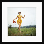 1970s Young Woman Wearing Colorful Print Dress Running In Field Carrying Red Straw Hat by Anonymous