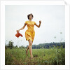 1970s Young Woman Wearing Colorful Print Dress Running In Field Carrying Red Straw Hat by Anonymous