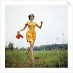1970s Young Woman Wearing Colorful Print Dress Running In Field Carrying Red Straw Hat by Anonymous