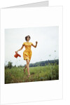 1970s Young Woman Wearing Colorful Print Dress Running In Field Carrying Red Straw Hat by Anonymous