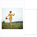 1970s Young Woman Wearing Colorful Print Dress Running In Field Carrying Red Straw Hat by Anonymous