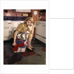 1960s Weary Dejected Woman Housewife Homemaker Sitting On Full Laundry Basket In Kitchen by Anonymous