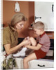1970s Mother Giving First-Aid Putting Band-Aid On Knee Of Son In Kitchen by Anonymous