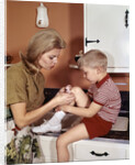1970s Mother Giving First-Aid Putting Band-Aid On Knee Of Son In Kitchen by Anonymous