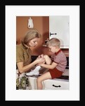 1970s Mother Giving First-Aid Putting Band-Aid On Knee Of Son In Kitchen by Anonymous