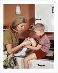 1970s Mother Giving First-Aid Putting Band-Aid On Knee Of Son In Kitchen by Anonymous